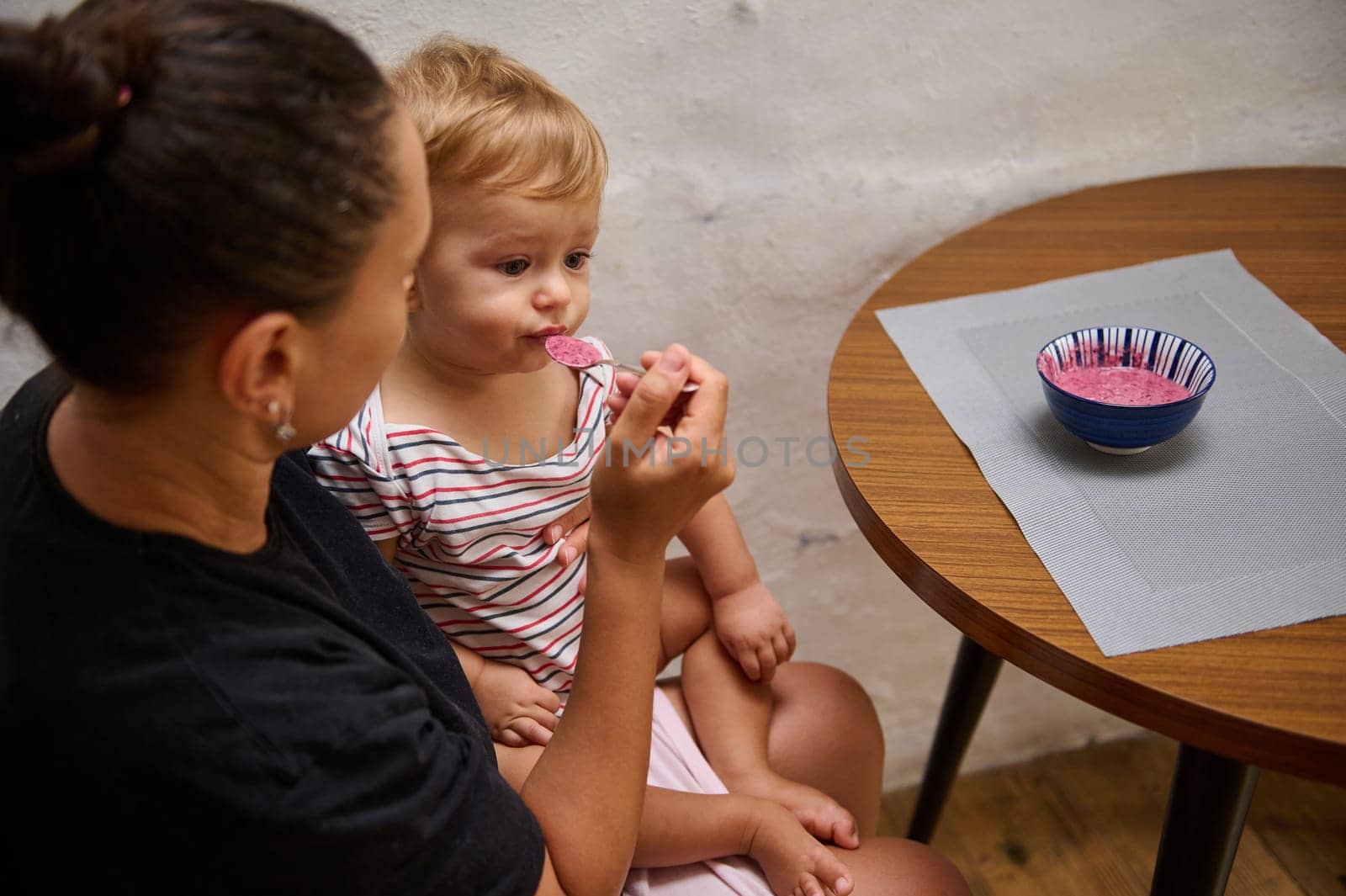 A mother lovingly feeds her baby with a spoonful of homemade fruit puree. The cozy home setting reflects nurturing and care, highlighting the bond between parent and child during mealtime.