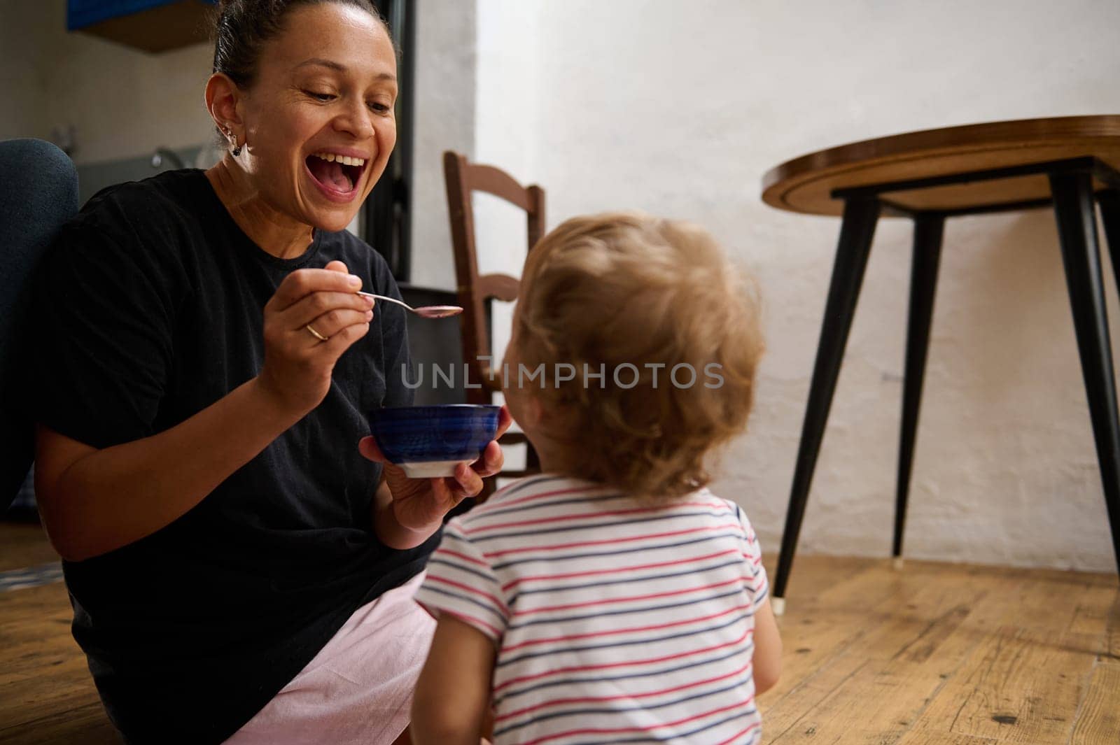 A joyful mother engages with her toddler during mealtime on the kitchen floor, sharing moments of laughter and bonding. The scene captures love, warmth, and family togetherness.