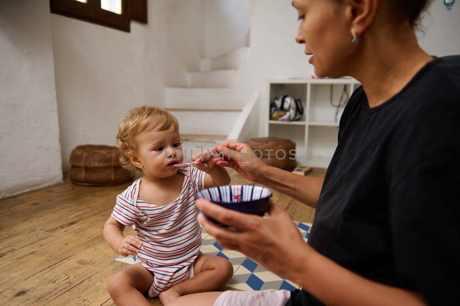 Mother feeds her toddler daughter with care and tenderness, sitting in a warm home setting. The child is enjoying a meal, showcasing a loving bond. The scene radiates comfort and warmth.