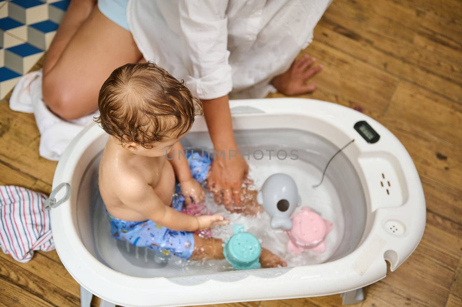 A mother assists her toddler during bath time, surrounded by colorful toys in a small tub, creating a joyful and nurturing scene. The wooden floor adds a cozy touch.