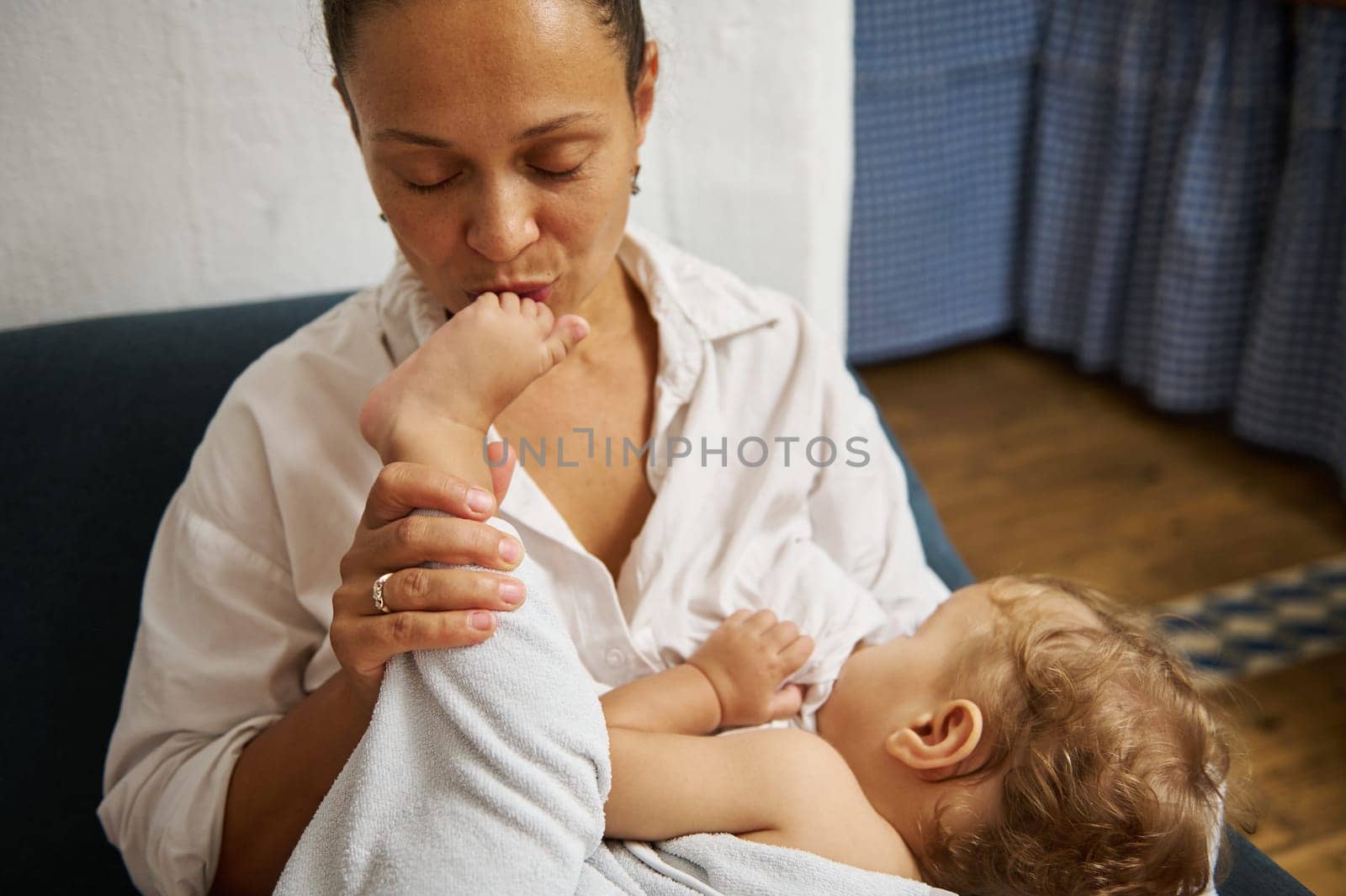 A mother expresses affection by gently kissing her baby's foot while cradling the child in a cozy setting, emphasizing warmth and love. The scene captures family bonding and tenderness.