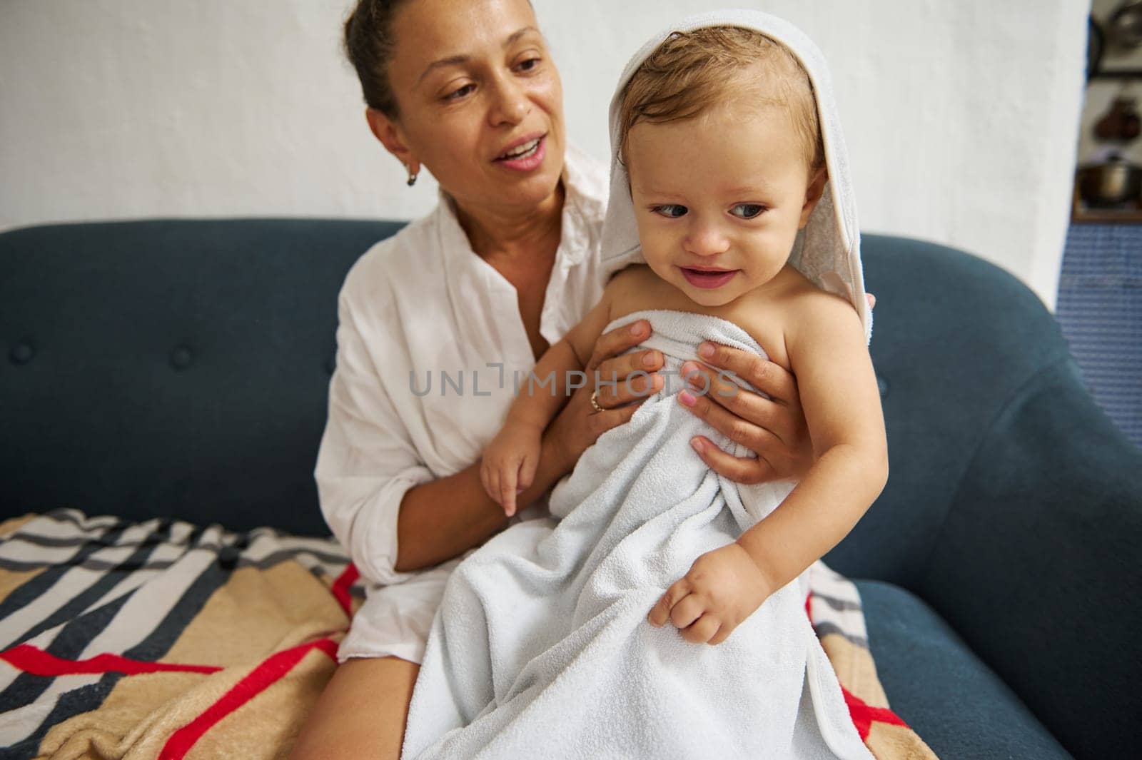 A joyful mother dries her smiling toddler with a soft towel on the couch, capturing a warm and tender moment of caregiving. The child looks content and playful after a bath, wrapped in comfort.