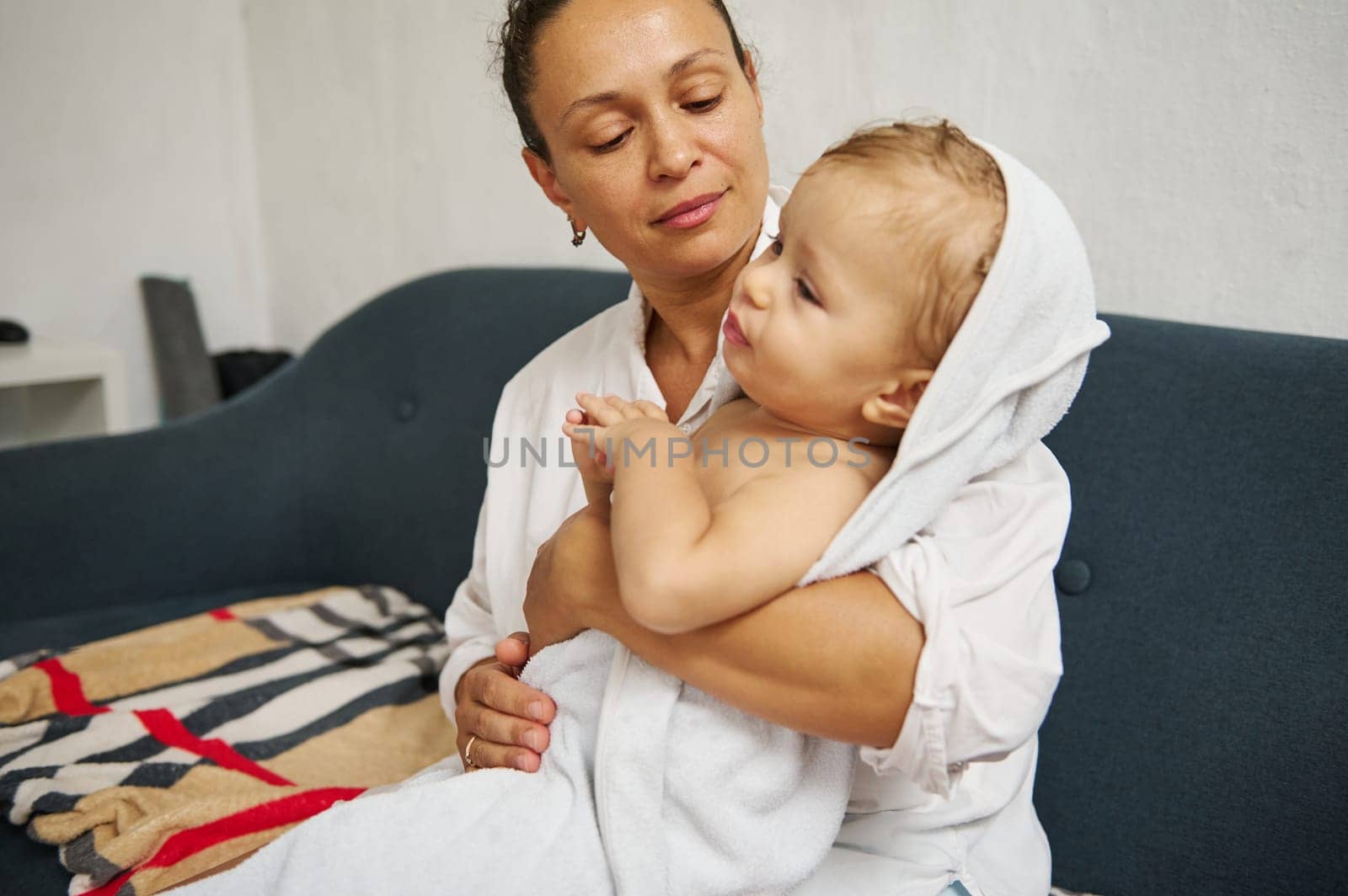 A caring mother holds her baby wrapped in a towel, sitting on a sofa. The scene portrays warmth, love, and comfort in a home setting, highlighting familial bonds and tenderness.