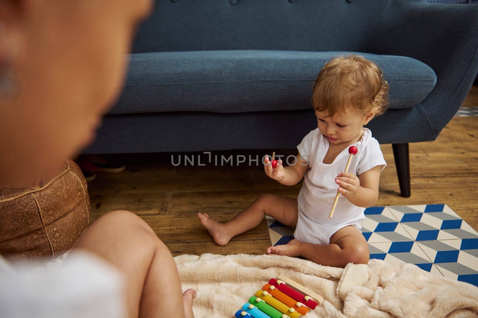A curious toddler in a white onesie plays with a colorful xylophone in a cozy living room setting, exploring sounds and textures.