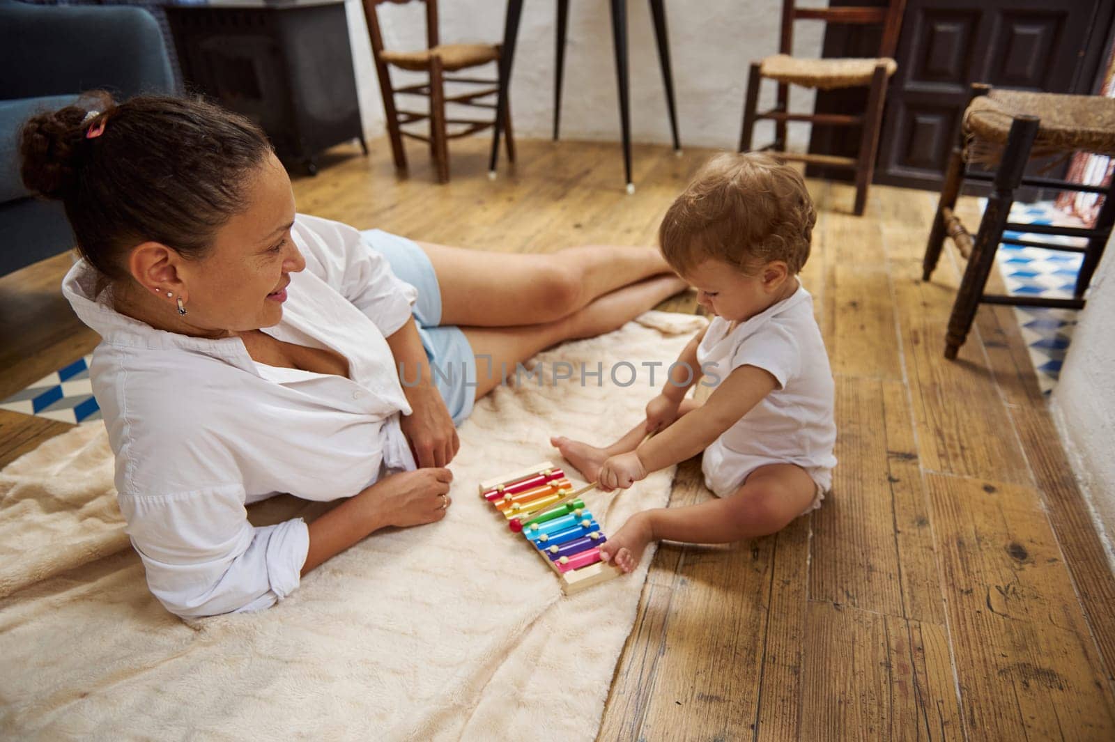 A mother and her baby enjoy quality time together, playing with a colorful xylophone on a wooden floor. The indoor setting adds warmth to this family bonding moment, filled with joy.