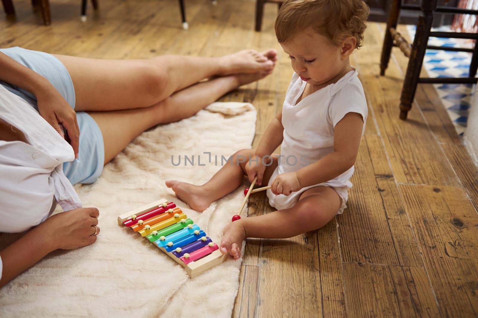 Cute toddler in a white onesie playing a colorful xylophone on a cozy blanket, next to a relaxed parent, creating a warm family atmosphere.