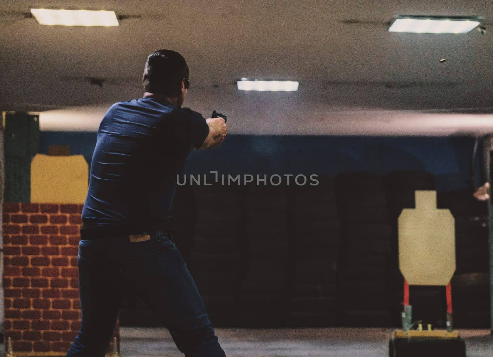 The image shows a person in a shooting range. The person is standing and aiming a handgun at a target downrange. The background includes typical shooting range elements such as an indoor setup with target silhouettes and protective barriers.
