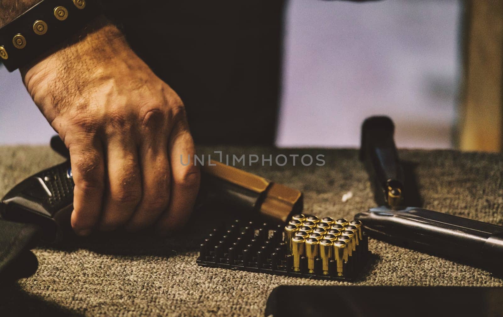 The image shows a person handling ammunition. There is a hand holding a handgun magazine, with a tray of bullets and parts of a firearm visible on a surface. The environment appears to be related to firearm handling or maintenance.