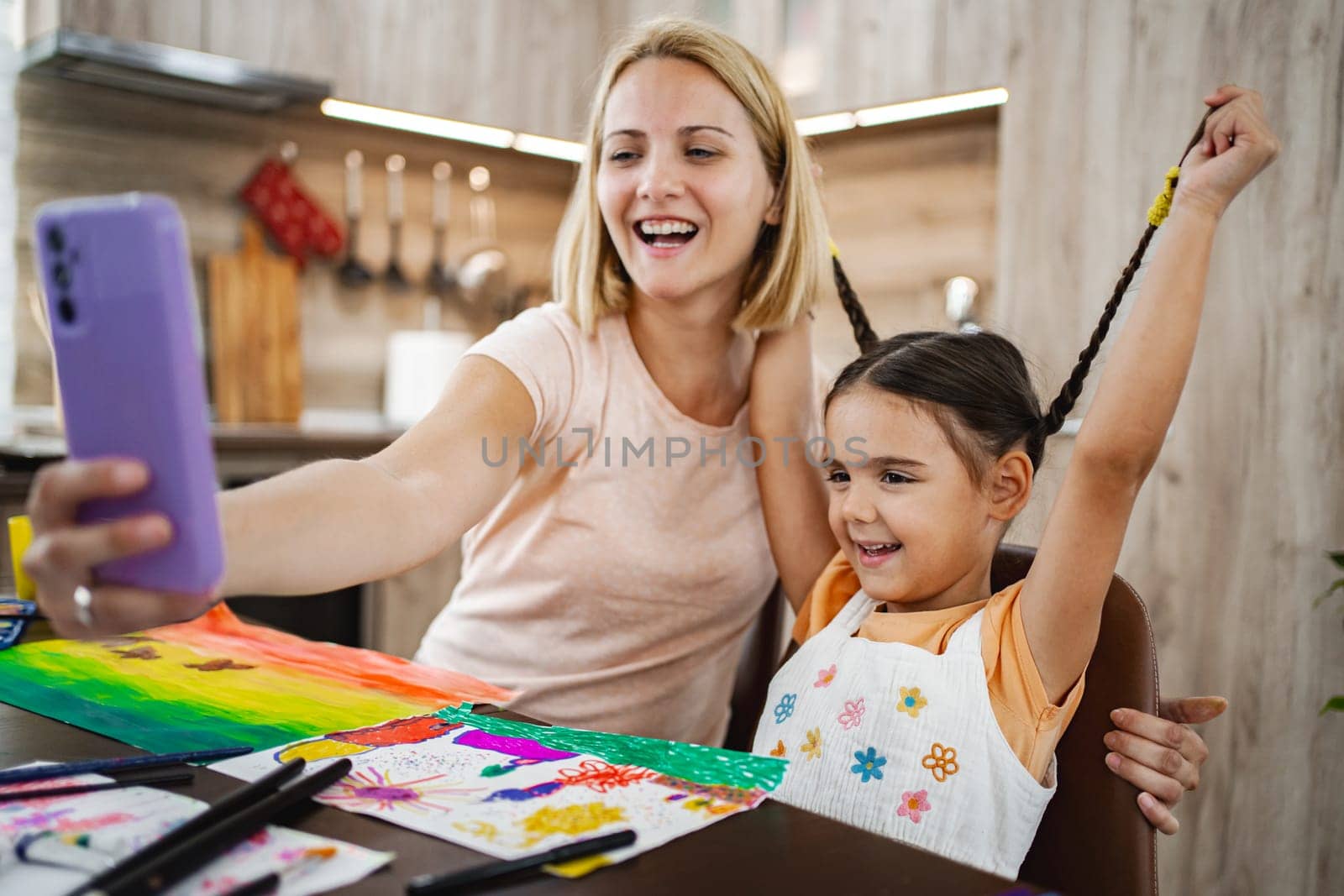 A mother and her daughter are laughing and taking a selfie together amid colorful art supplies at their home.