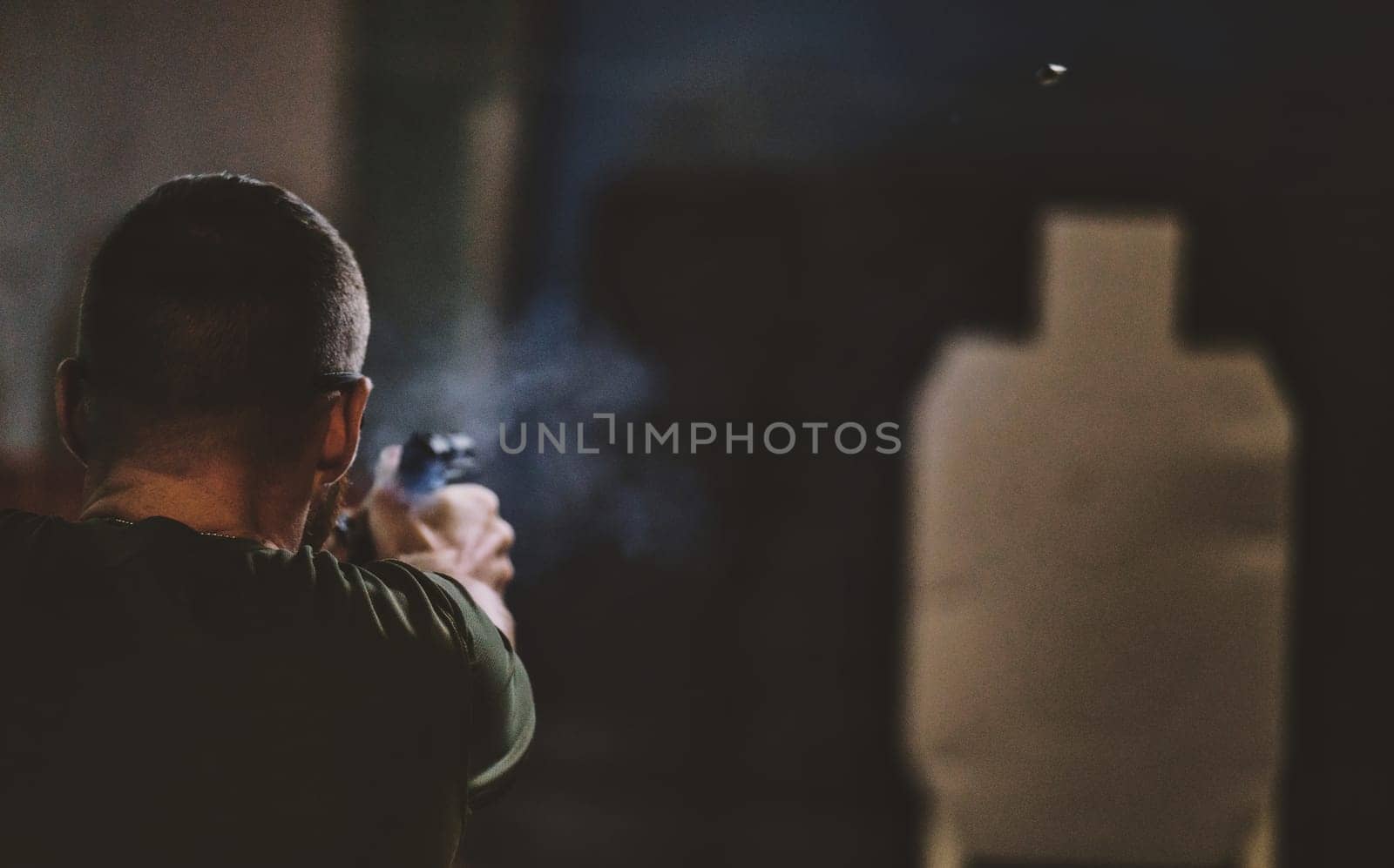 The image shows a person shooting a firearm at a shooting range. The person is aiming at a target silhouette. There is visible smoke near the firearm, likely from a recent shot. The angle of the image is from behind the person, focusing on the act of shooting and the target ahead.