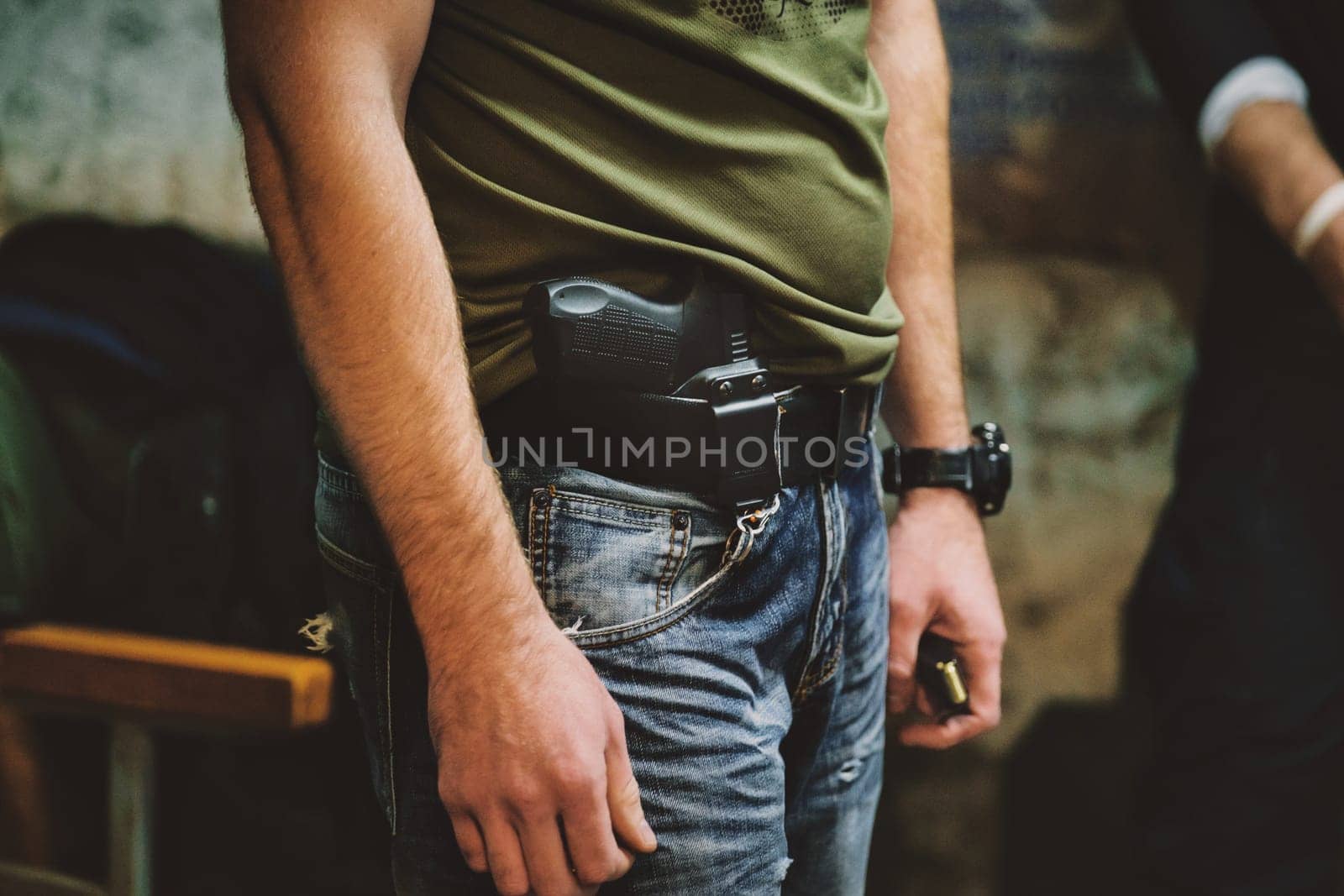 The image shows the midsection of a person wearing jeans and a green shirt. The person has a handgun holstered at their waist. They are also wearing a black wristwatch on their left wrist. The background is somewhat blurred but appears to consist of a bench and possibly some other indistinguishable objects or people.
