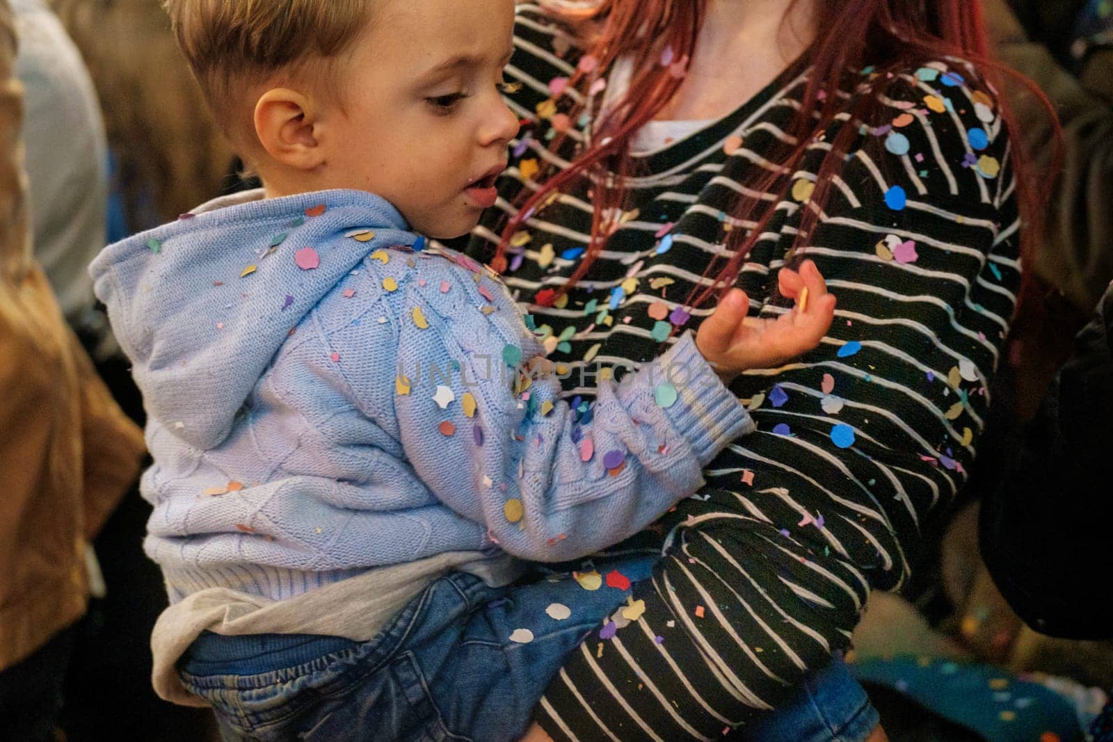 The image shows a young child being held by an adult. The child appears to be looking with curiosity at something on their hand. Both the child's blue jacket and the adult's clothing are speckled with colorful confetti, suggesting they may be at a festive event, such as a party or celebration where confetti was thrown. The setting seems to be indoors with other people nearby, indicated by the partial view of another person in the background.