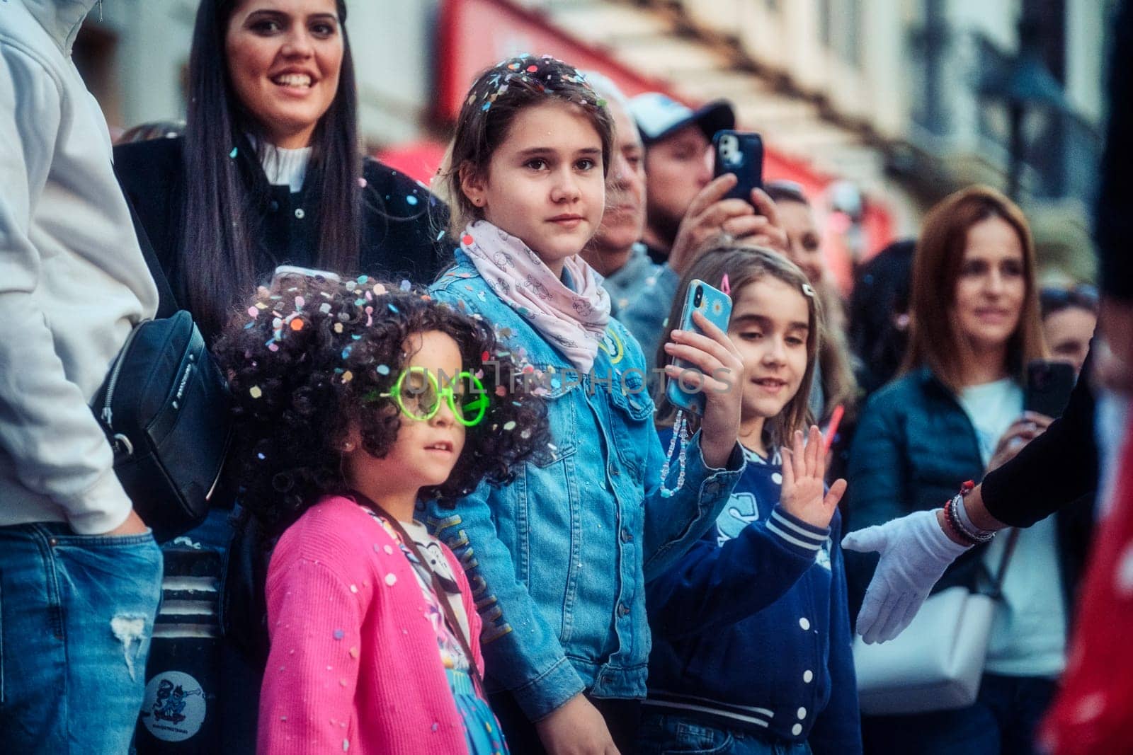 Enthusiastic crowd at an outdoor event with two focused girls in the forefront, one wearing quirky glasses and others capturing the moment on smartphones.