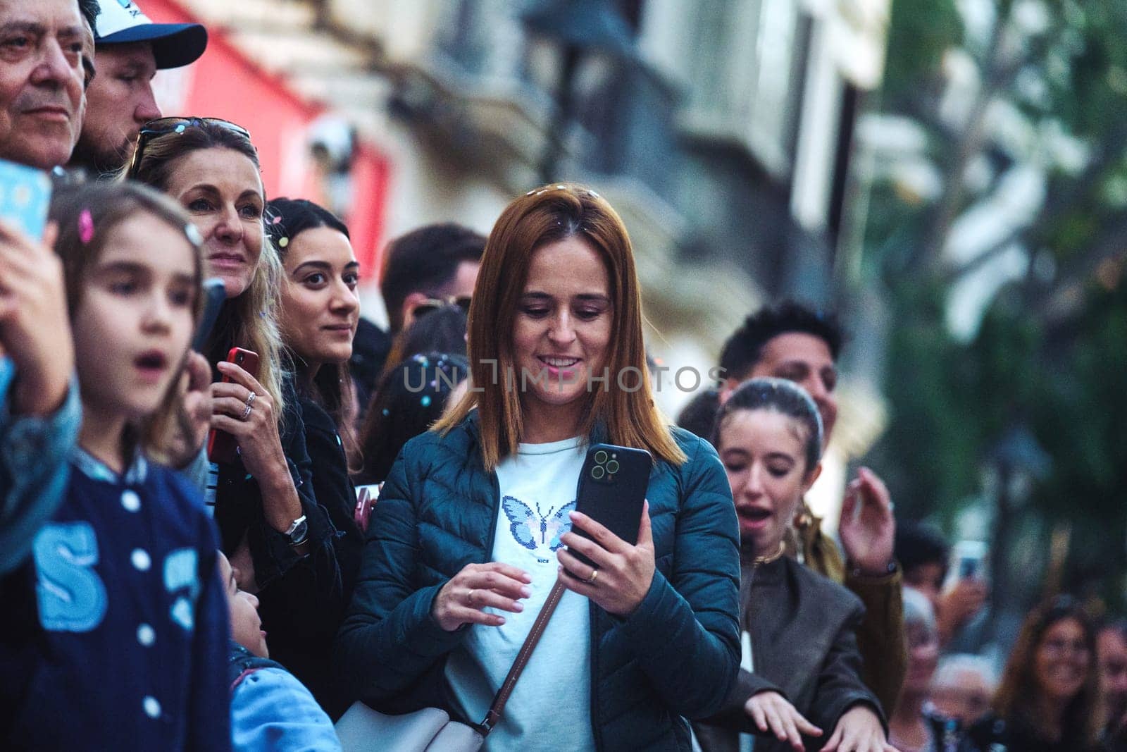 In this image, there is a crowd of people, and they appear to be focused on an event or something of interest not shown in the photo. At the forefront, there is a woman with reddish-brown hair who is looking down at her smartphone, which she is holding with both hands. People in the background have various expressions, and there are a couple of individuals who are also capturing the moment with their phones. The setting looks like a city street with buildings in the background, suggesting an urban outdoor event or gathering. The shallow depth of field places the emphasis on the woman with the phone.