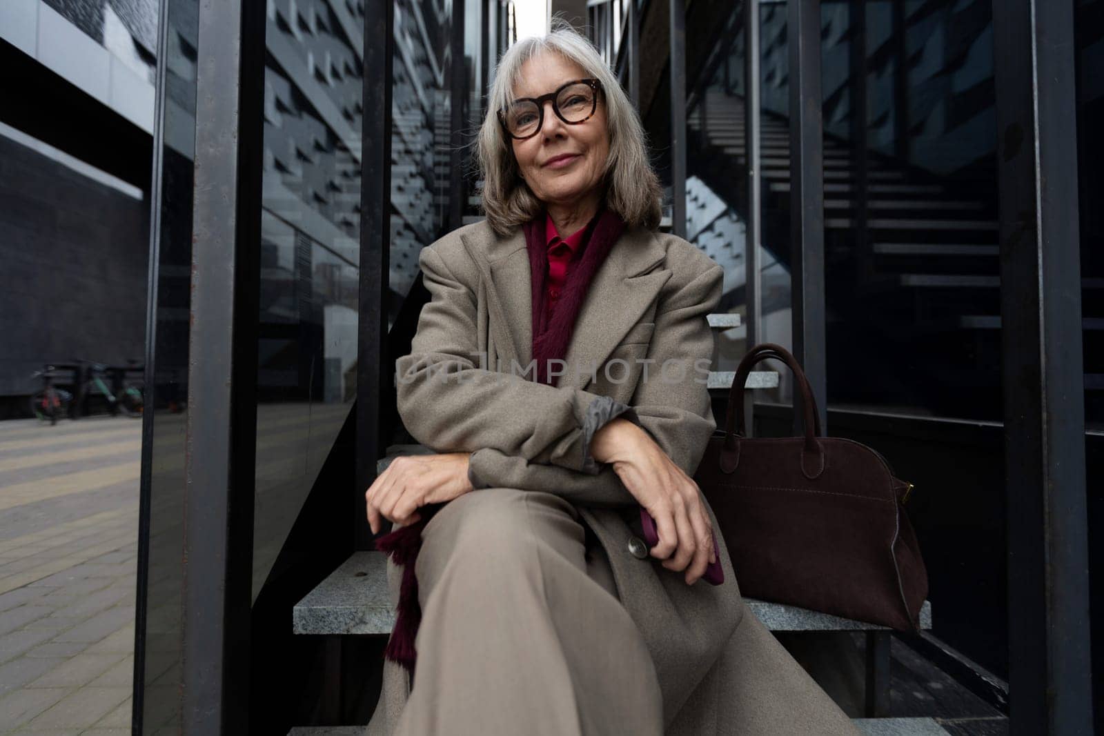 A middle-aged businesswoman with glasses relaxes on the steps outside a contemporary building. She wears a stylish outfit and conveys a sense of confidence and professionalism.