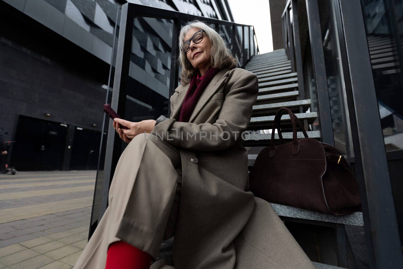 A mature businesswoman dressed in a stylish coat and vibrant red stockings sits on the stairs of a contemporary building, focused on her smartphone while enjoying the urban atmosphere.