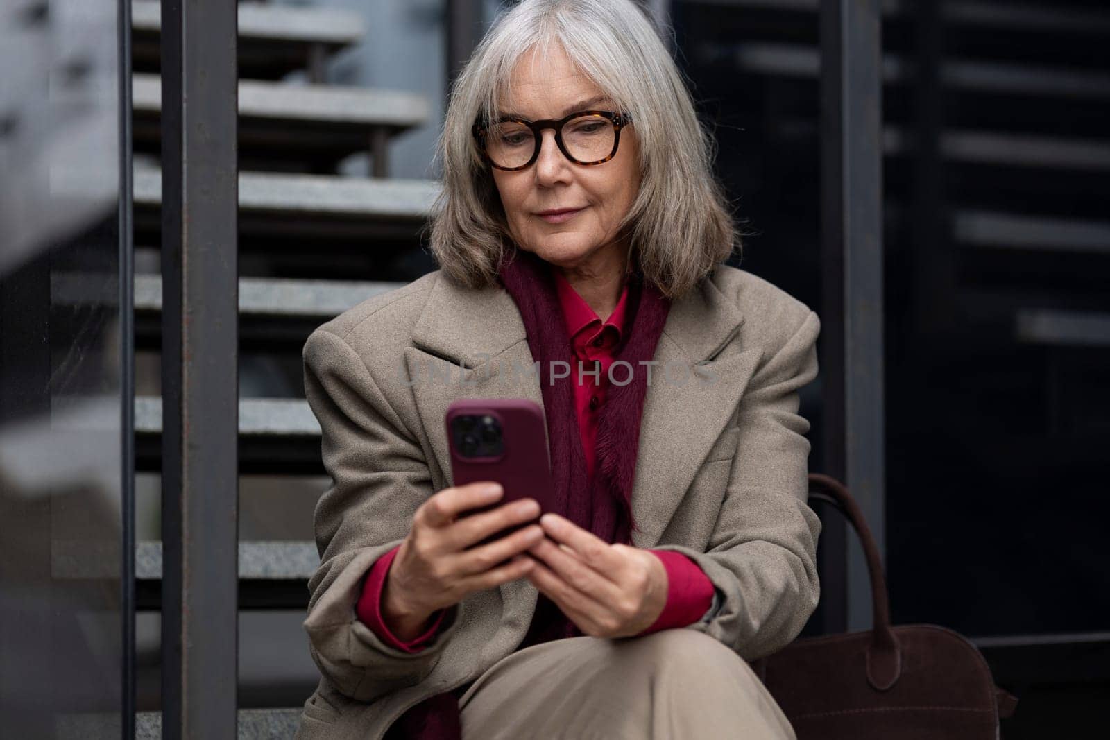 An elderly woman with gray hair is sitting on a set of steps outside, wearing a beige blazer over a red shirt. She is looking down at a smartphone in her hands.