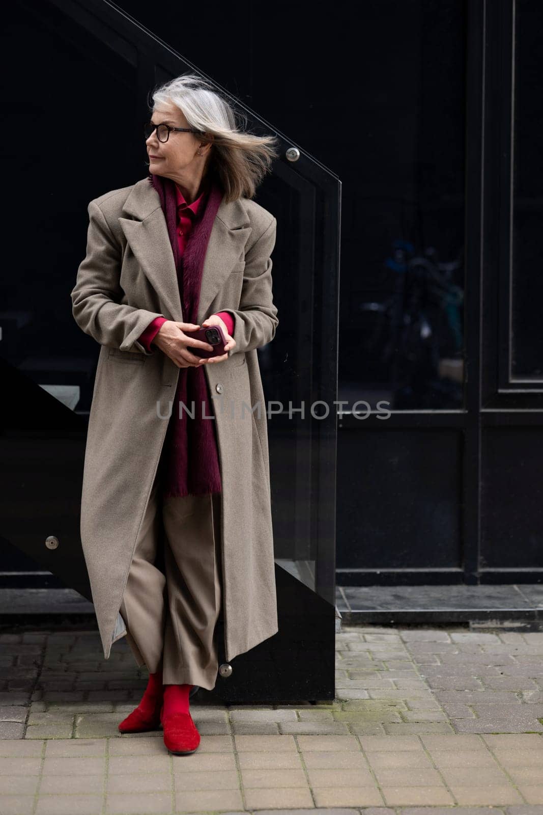 An elderly woman with gray hair wears a chic coat and red boots while standing against a dark glass building. The backdrop suggests a lively urban atmosphere in autumn.