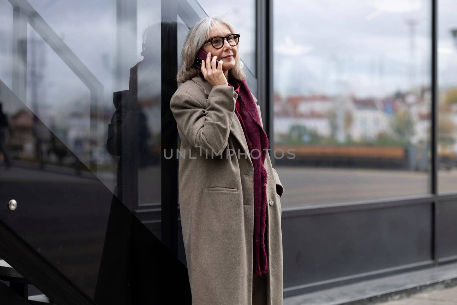 The older woman is casually leaning against a sleek glass wall, speaking cheerfully on her phone while dressed comfortably for the day.
