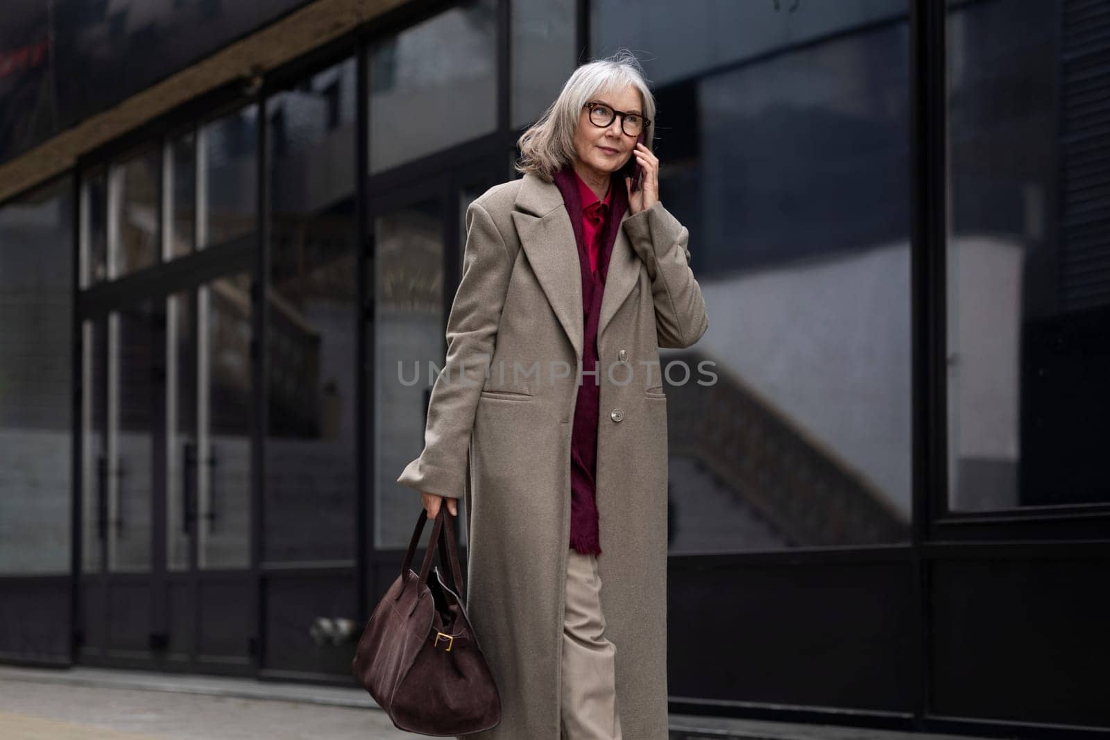 A senior businesswoman is walking confidently outdoors, talking on her phone. Her professional attire and focused demeanor reflect her dedication and engagement in the city s business environment.