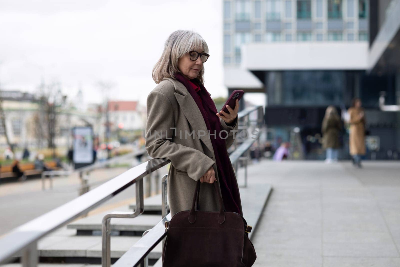 A senior businesswoman stands confidently outside a contemporary building, checking her smartphone while dressed in a stylish coat. The cool atmosphere suggests a brisk day.