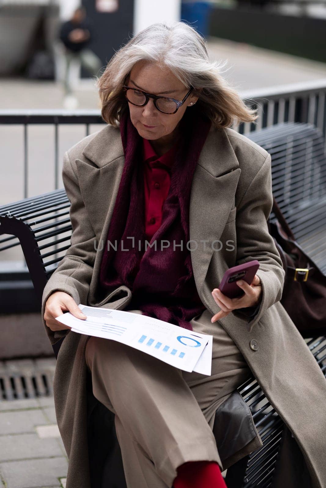 A thoughtful woman examines paperwork while seated on a bench in an urban setting. She holds a phone in her other hand and wears a stylish coat.