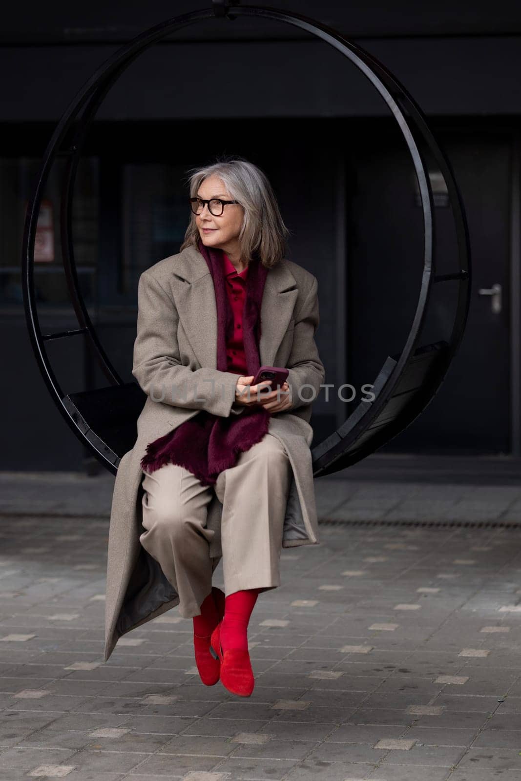 A stylish elderly woman dressed in a beige coat and red scarf sits gracefully on a circular swing, holding a phone. The modern urban backdrop adds to her chic appearance.