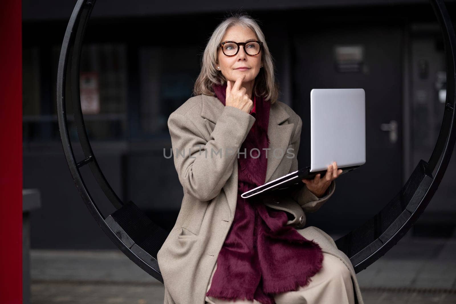 An older woman thoughtfully holds her laptop while sitting on a modern circular seat. She wears a fashionable coat and looks contemplative in an outdoor urban environment.