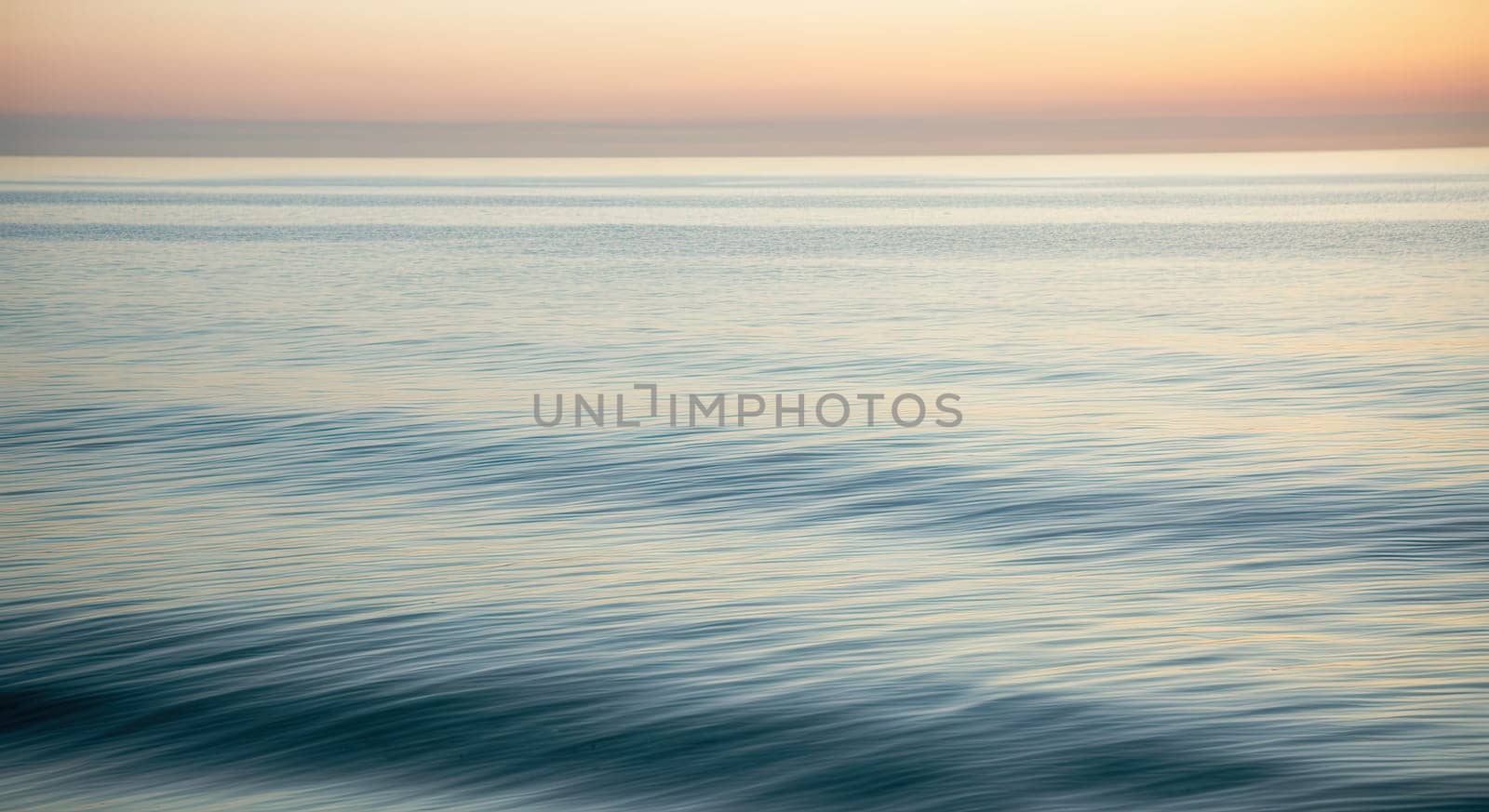 Serene ocean waves ripple under a tranquil sunset, showcasing soft pastel hues of the sky meeting the reflective sea. ideal as a calming background for design projects or prints, emphasizing peace and natural beauty.