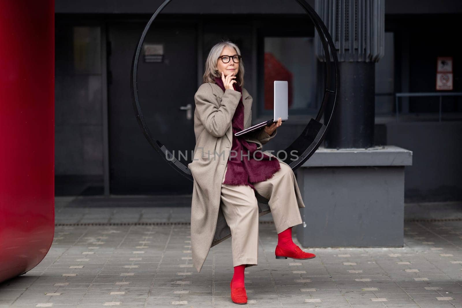 An elderly businesswoman, elegantly dressed in layers, engages with her laptop while seated on a modern circular bench in an urban environment, showcasing a blend of professionalism and creativity.