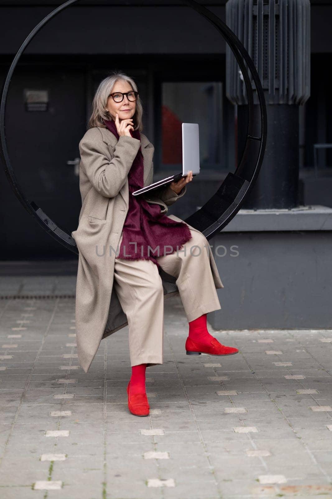 An elderly woman with a fashionable appearance sits thoughtfully in a circular outdoor seat, engaging with her laptop while enjoying the urban atmosphere on a sunny day.