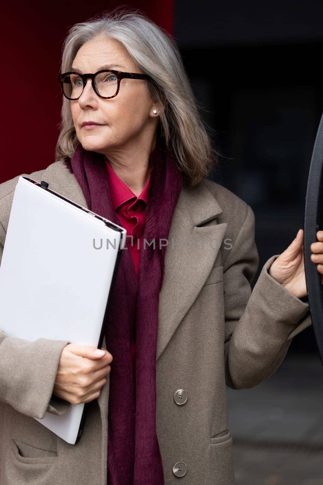 A senior businesswoman with gray hair stands outside, dressed in a tailored coat and scarf. She holds a clipboard, looking thoughtfully towards her surroundings, embodying professionalism.