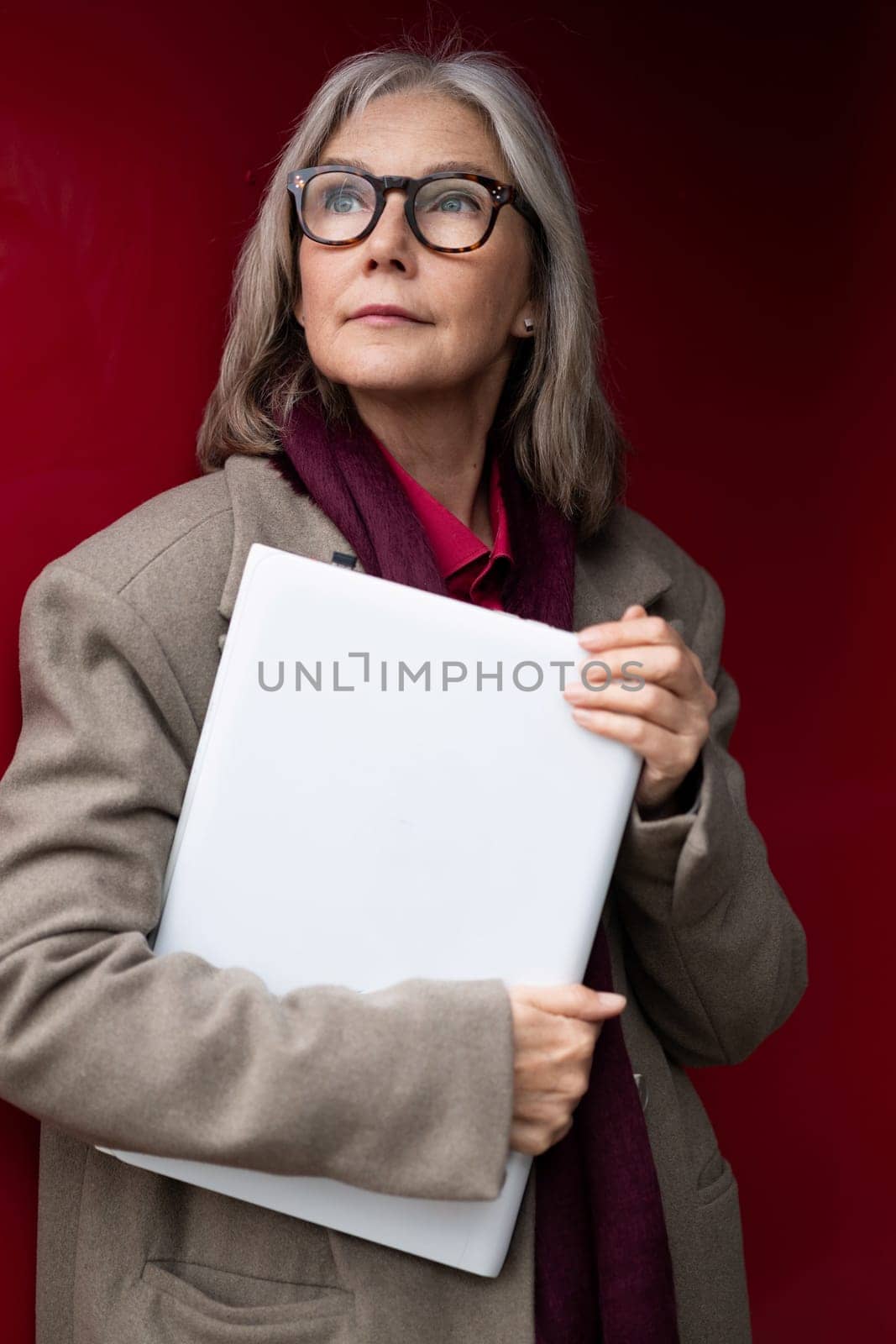 An elderly woman with stylish glasses and gray hair stands with a confident posture, holding a folder close to her chest against a vibrant red background.