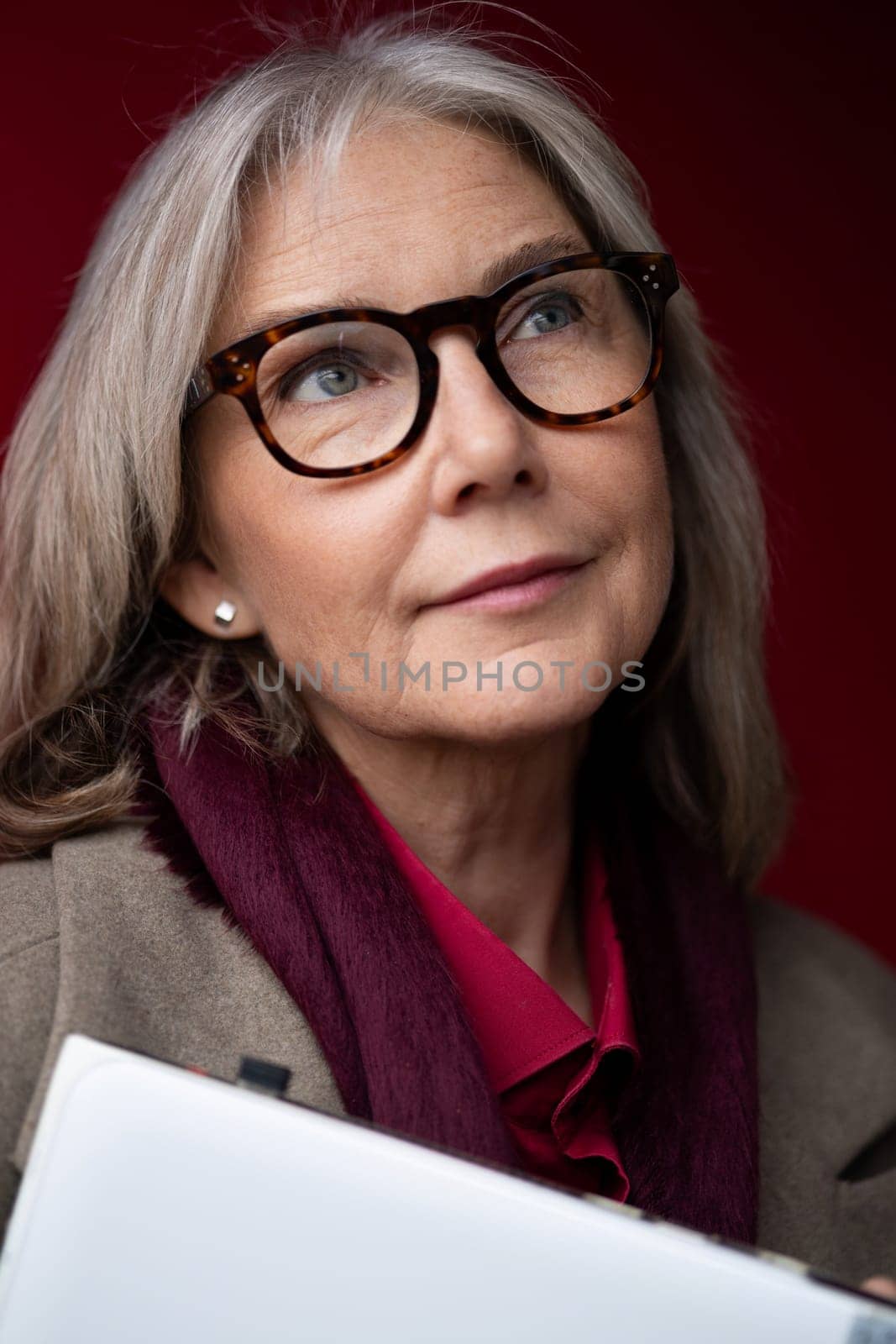 The older woman gazes upward with a pensive expression, wearing glasses and a scarf. She holds a notepad, suggesting contemplation or planning in a cozy setting.
