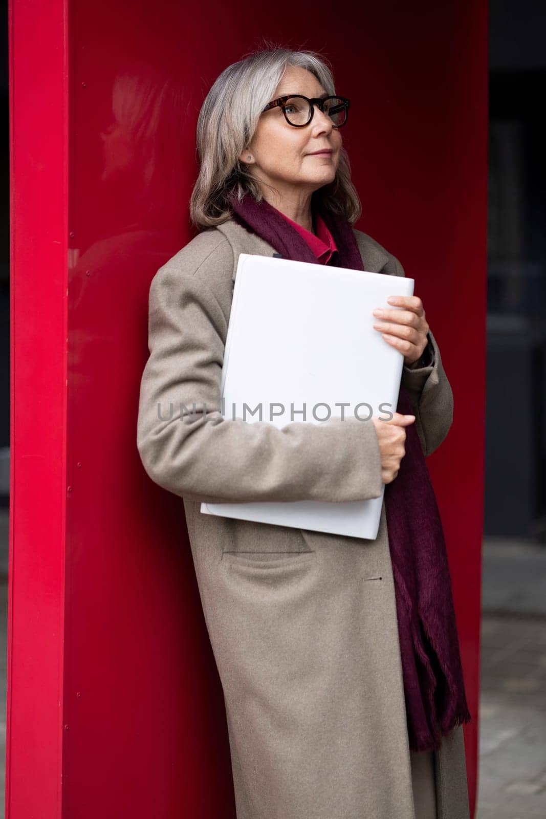 The older woman leans against a vivid red wall, dressed in a beige coat with a maroon scarf. She holds a white folder close, appearing reflective and engaged in deep thought.