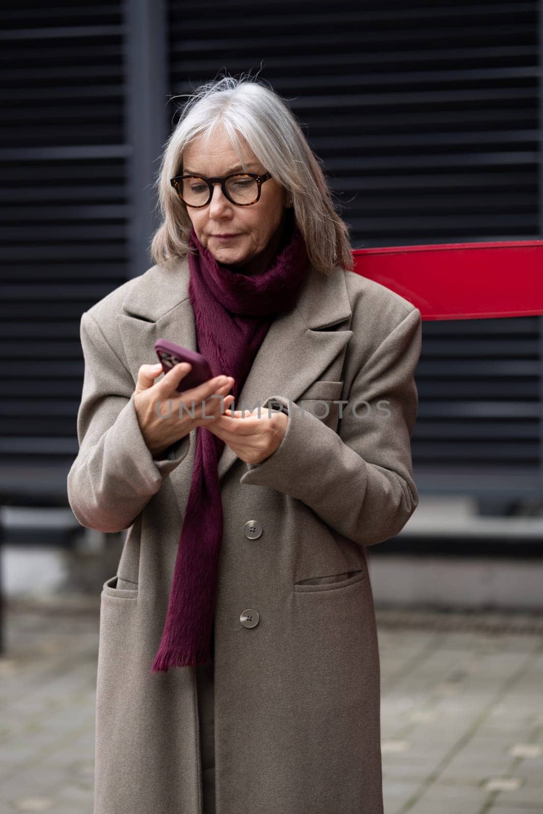 An older woman in a stylish coat and scarf stands outside, intently checking her phone while surrounded by a modern urban environment on a cool day.