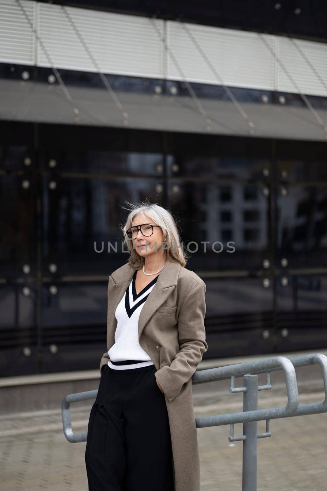 The senior businesswoman exudes confidence as she stands outside a sleek, contemporary building. Dressed in a chic outfit, she gazes thoughtfully, embodying professionalism.