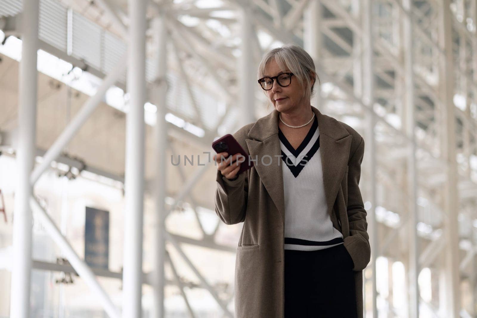 A middle-aged Caucasian businesswoman stands in a contemporary office space, absorbed in her smartphone, dressed elegantly in neutral tones against a sleek backdrop.