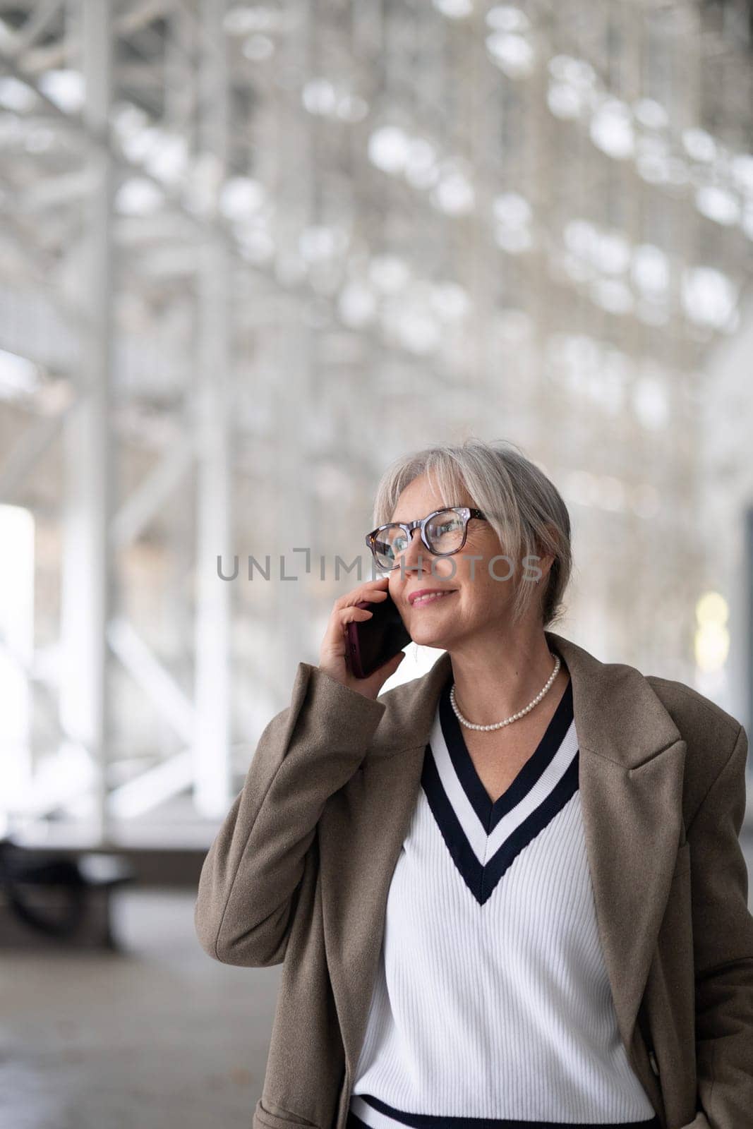 The elderly businesswoman, dressed elegantly in a sweater and blazer, smiles while conversing on her phone in a bright, contemporary office space, showcasing her confidence and poise.