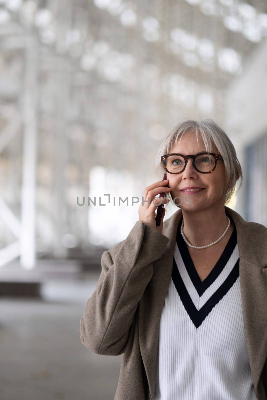 A senior businesswoman enjoys a phone conversation while strolling through a contemporary outdoor area. She appears confident and engaged, dressed smartly for the day.