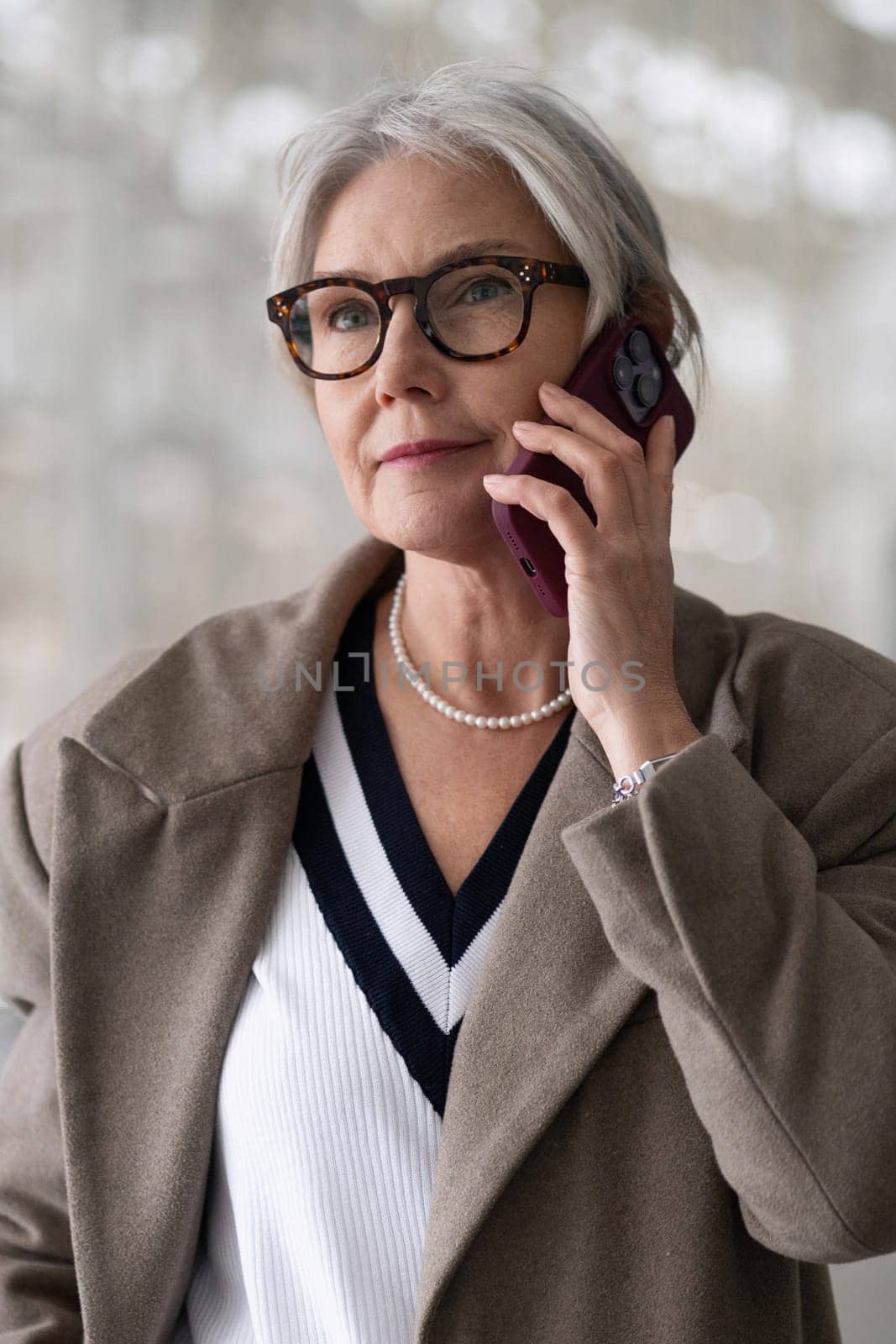 An older woman engages in a conversation on her phone, dressed elegantly in a coat and pearl necklace, expressing a moment of thoughtfulness and sophistication in her surroundings.