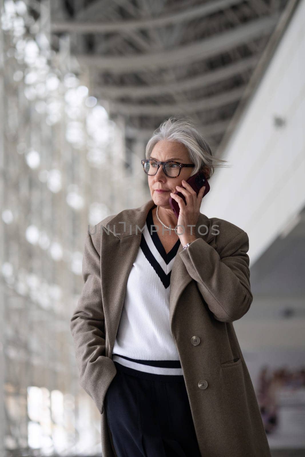 A businesswoman with gray hair and glasses walks confidently through a contemporary indoor environment, speaking on her mobile phone and dressed in a fashionable outfit.