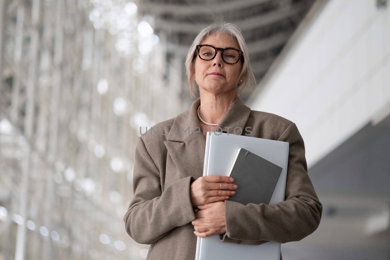 A middle-aged businesswoman stands confidently in a contemporary office setting, holding documents and a laptop. She appears focused and professional amid sleek architecture.