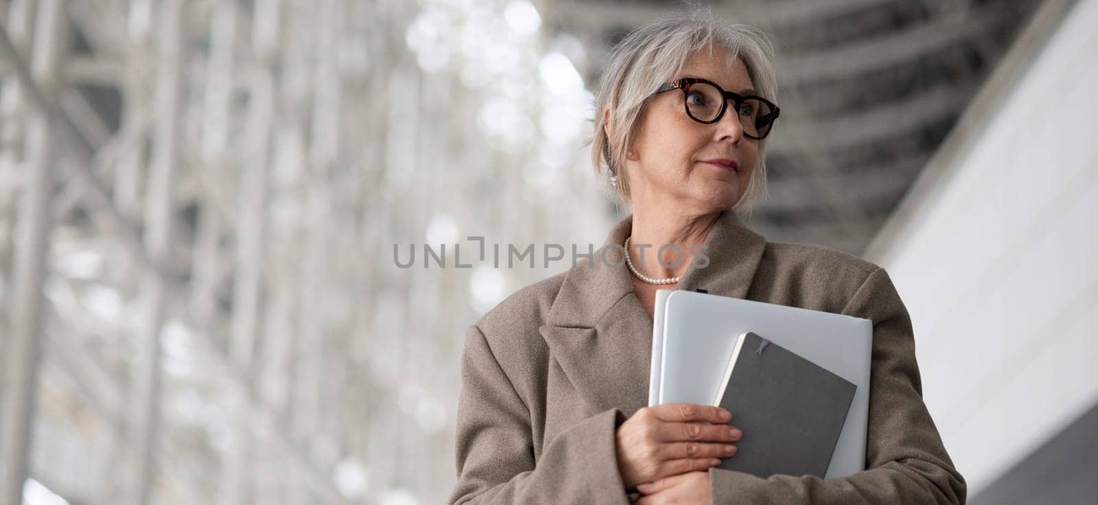 The middle-aged businesswoman, dressed in a professional outfit, thoughtfully gazes ahead while holding a laptop and notebook in a contemporary office environment.