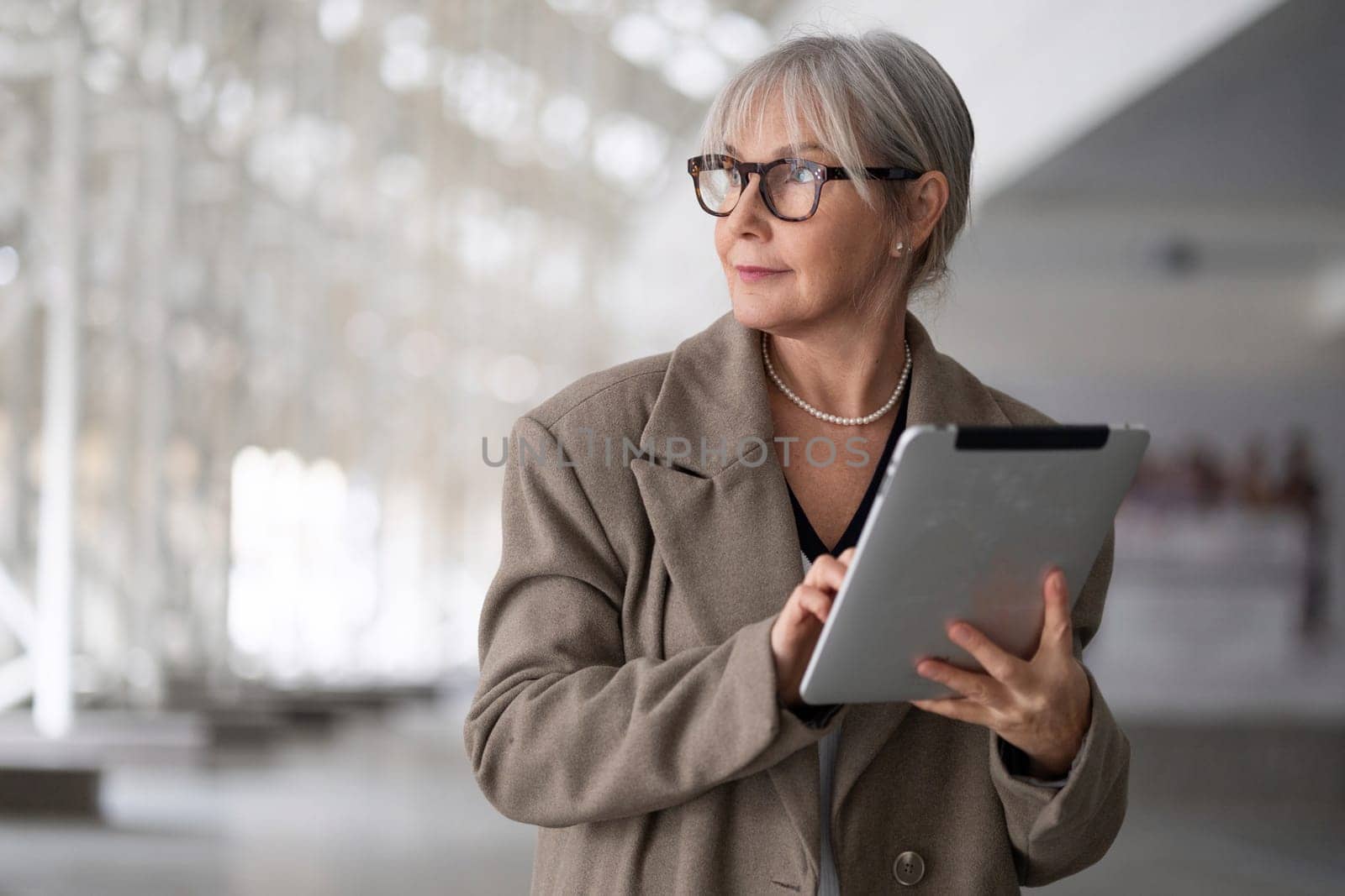 A middle-aged businesswoman with glasses holds a tablet, appearing focused and engaged in her work inside a contemporary office space. Her attire reflects a professional setting.