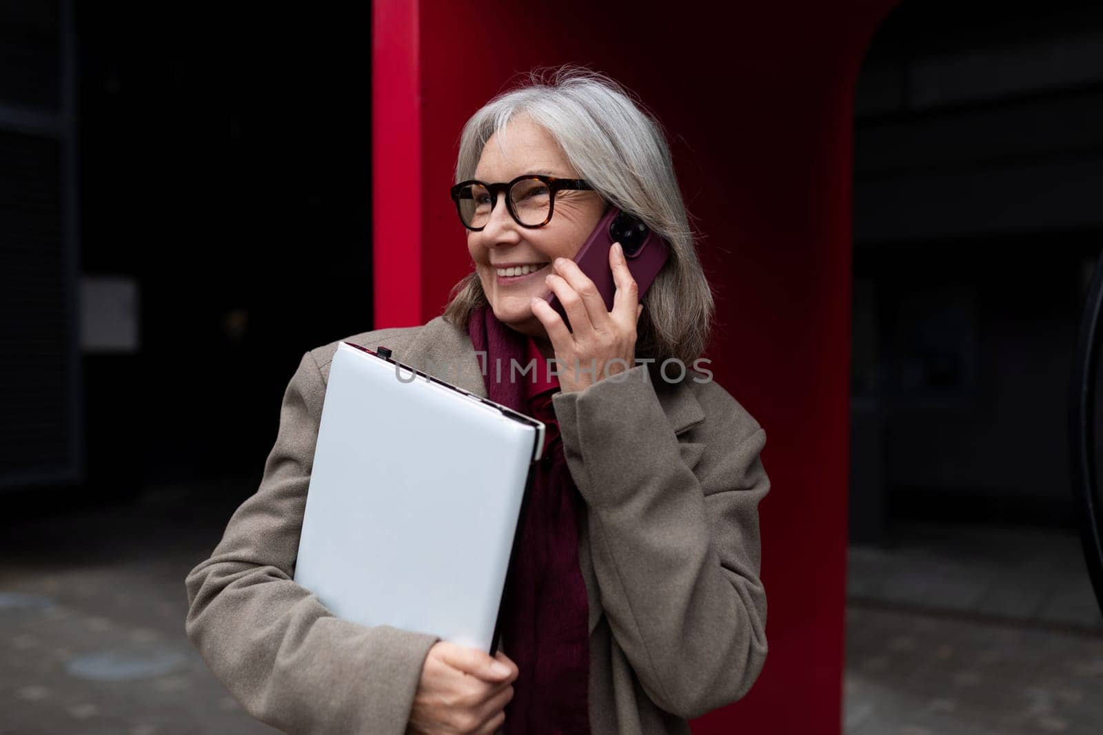 A middle-aged Caucasian businesswoman enjoys a cheerful phone conversation while holding a folder. She stands outside an office building, radiating confidence and approachability.