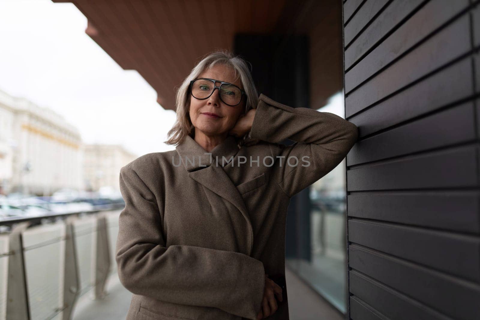 An elderly businesswoman leans against a stylish exterior wall outside her office. She wears glasses and a tailored coat, exuding confidence and professionalism in a bustling urban area.