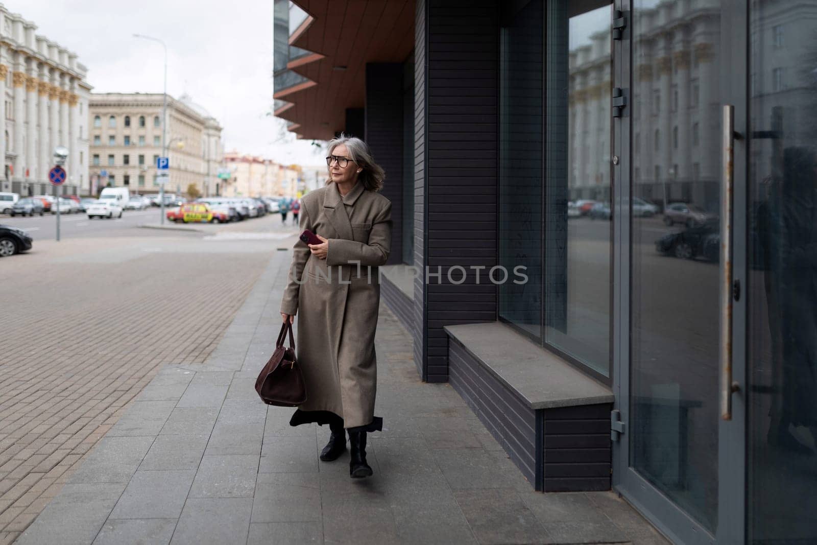 A confident elderly businesswoman strolls along a busy city street, wearing a stylish coat and carrying a handbag, displaying poise and professionalism.