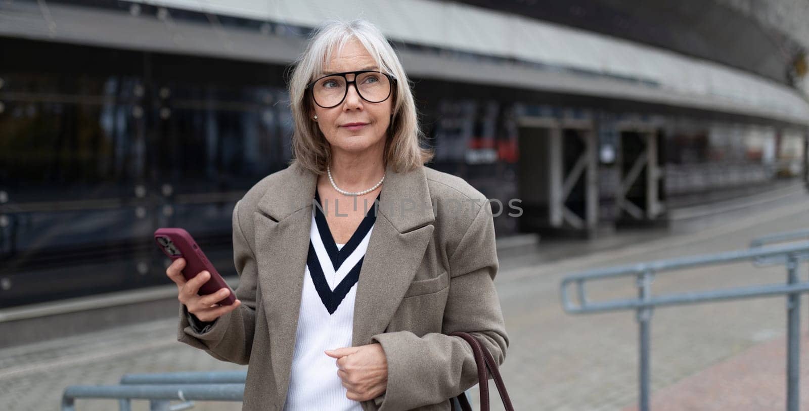 An elderly businesswoman in a stylish outfit holds her phone as she stands outside a modern office building, enjoying the sunny day while looking thoughtful.