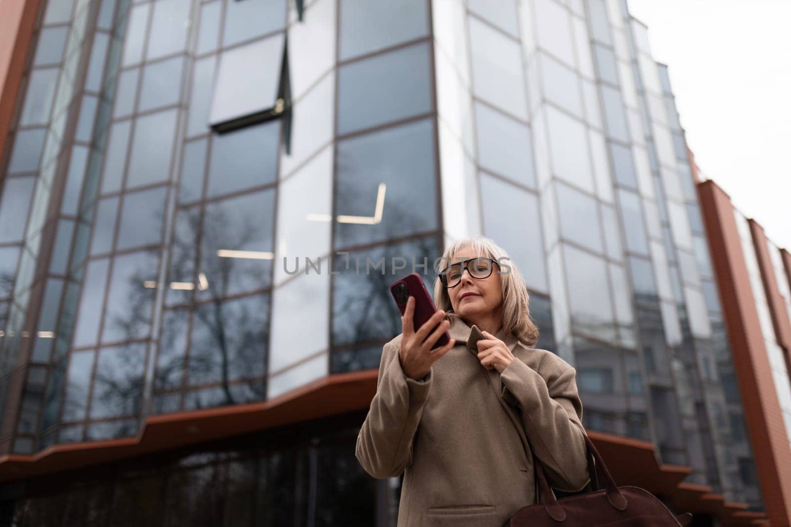 A senior businesswoman stands outside a contemporary office building, focused on her smartphone. She wears a stylish coat and holds a bag, appearing engaged in her task.